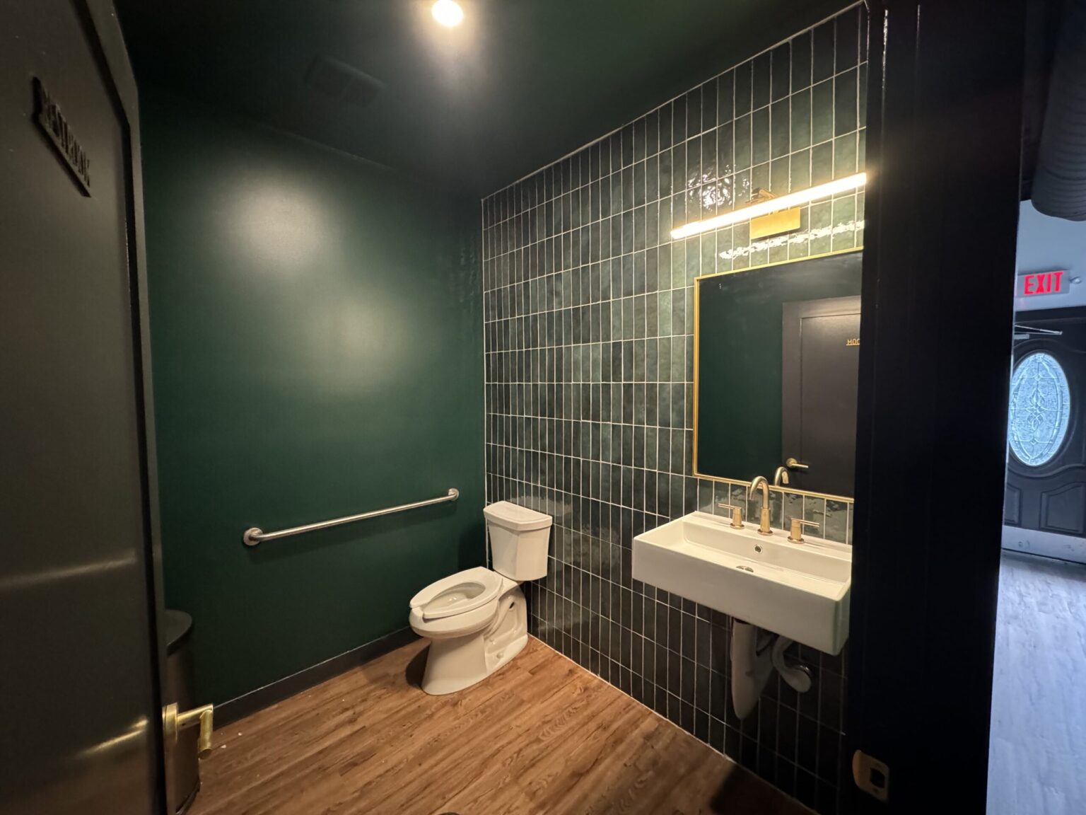 Office restroom in a Saline commercial build-out with deep green walls, stacked green tile, a white wall-mounted sink, a brass-framed mirror and a grab bar beside the toilet