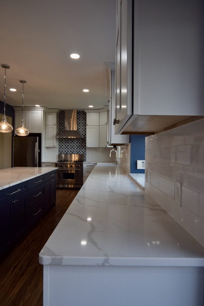 Long run of white veined counters and white cabinets beside a navy island, with wood-look plank flooring running the length of the kitchen