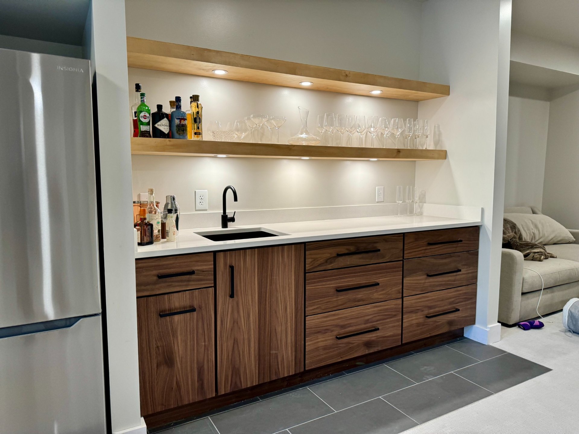 A finished basement with a wet bar, floating wood shelves, bottles, and under-cabinet lighting