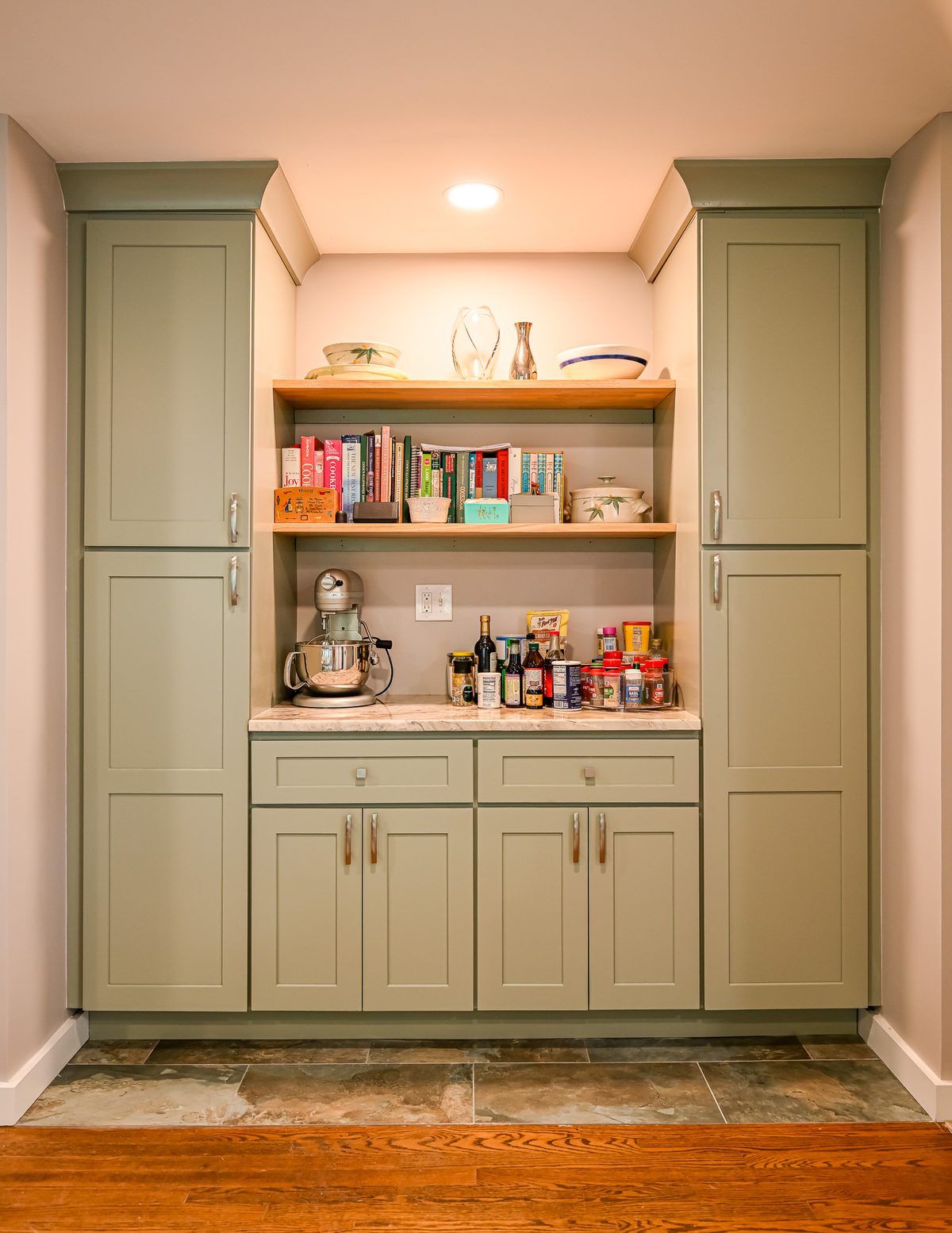 Close-up of the sage green built-in pantry with two open wood shelves holding cookbooks and decorative bowls, a marble countertop with a KitchenAid stand mixer and spice collection, and tall shaker cabinets flanking each side