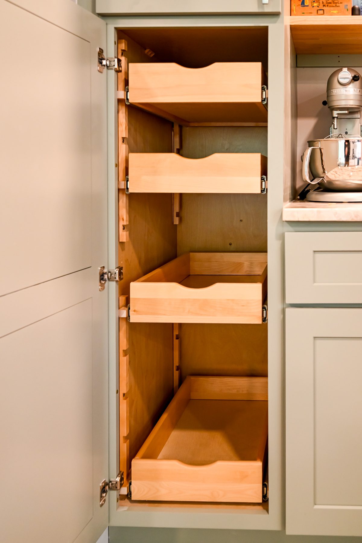 Interior of the sage green pantry cabinet showing four pull-out wooden shelf trays with scalloped front edges, mounted on full-extension soft-close drawer slides