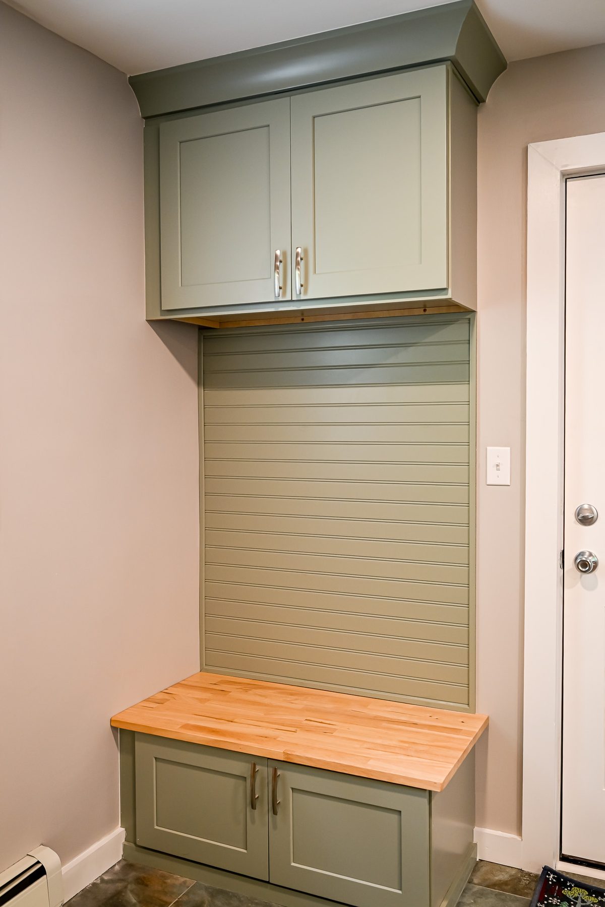Close-up of the mudroom bench showing sage green shaker cabinet base with butcher block top, a slatwall organizer panel in matching sage green, and an upper storage cabinet with crown molding detail
