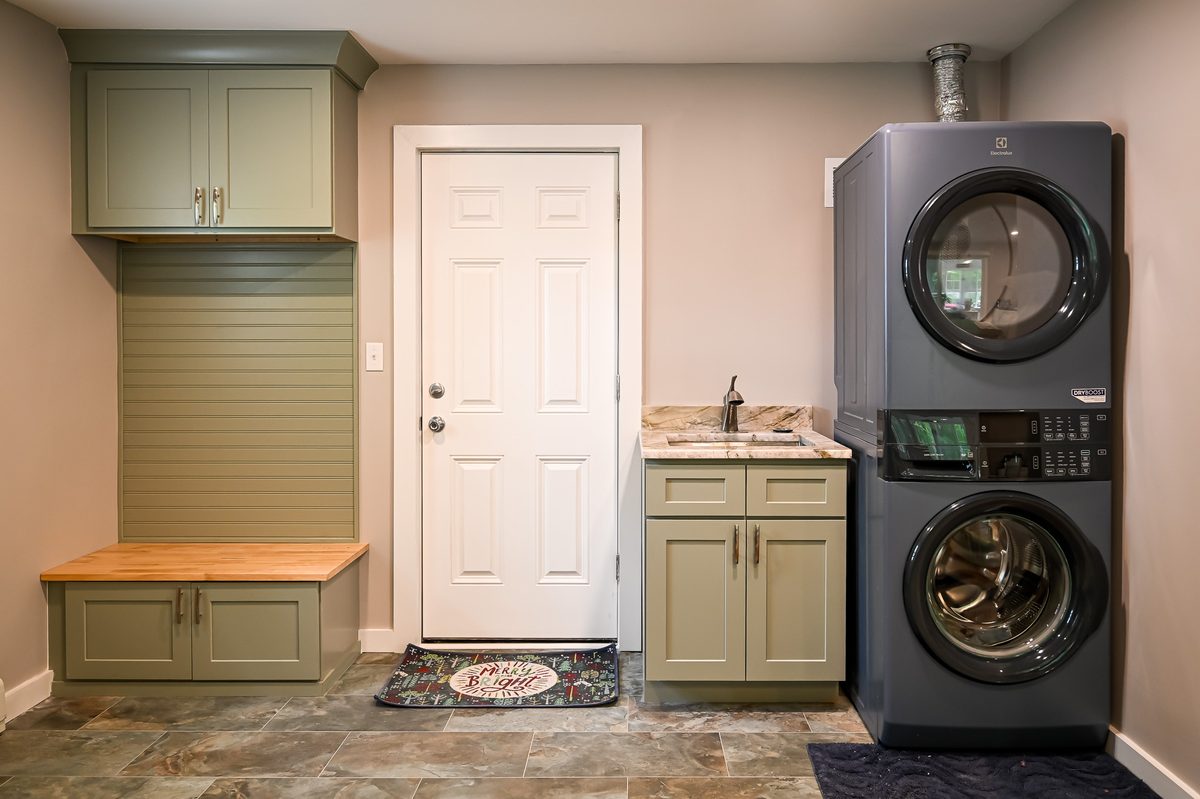 Mudroom entry area with a sage green bench cabinet and butcher block seat, slatwall panel for hooks, an upper storage cabinet, a utility sink with marble countertop, and a stacked Electrolux washer and dryer unit next to the exterior door