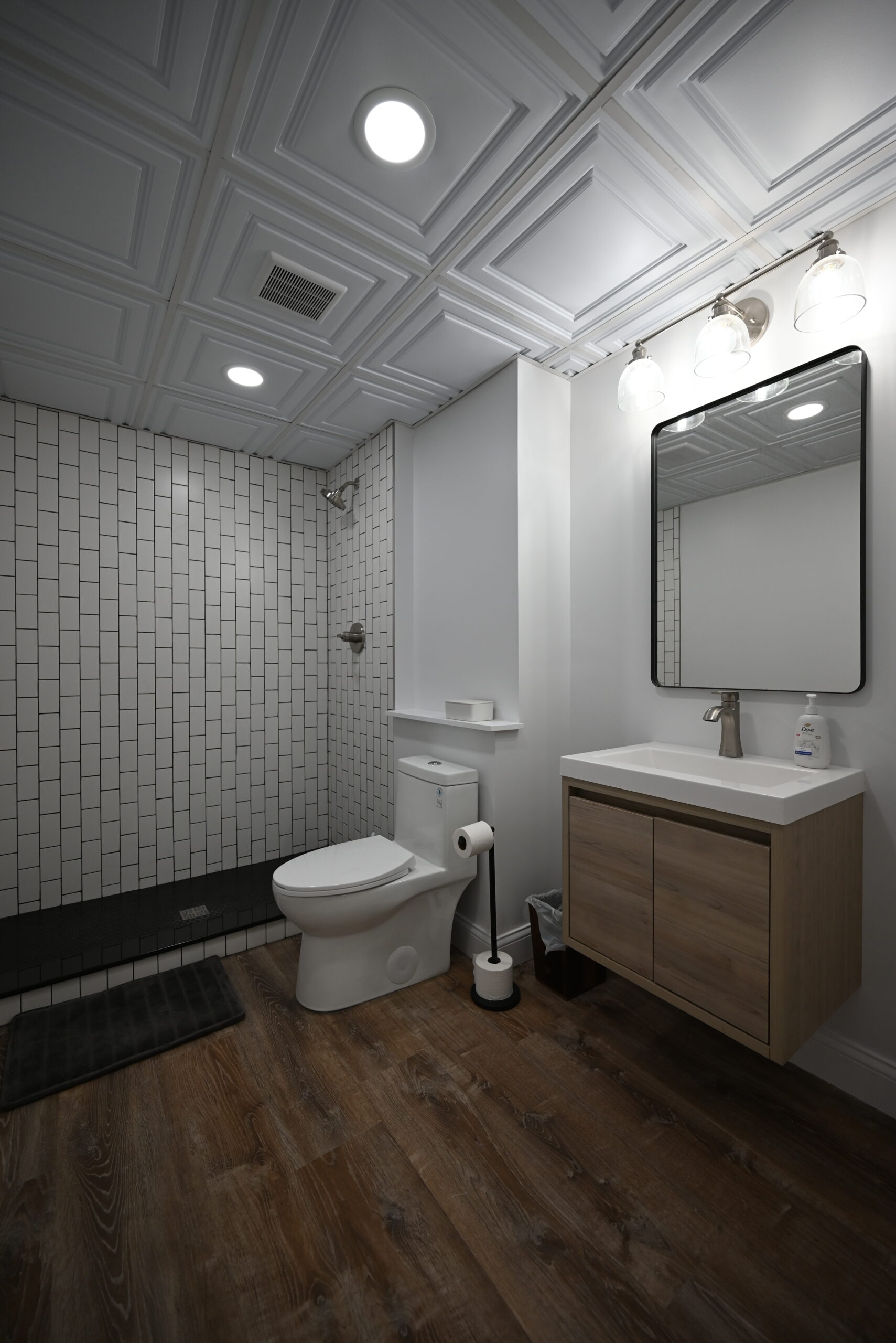 Basement bathroom showing walk-in shower, floating wood-tone vanity, black-framed mirror, globe light fixtures, and decorative ceiling tiles