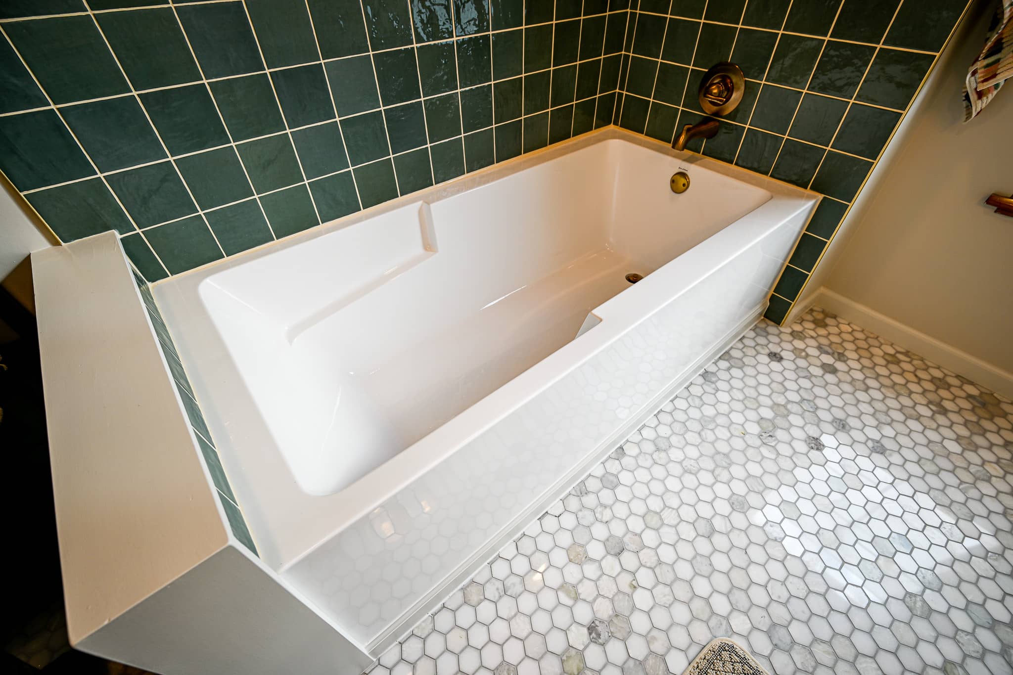 White alcove soaking tub set into green tile above a white marble hexagon floor in Dexter