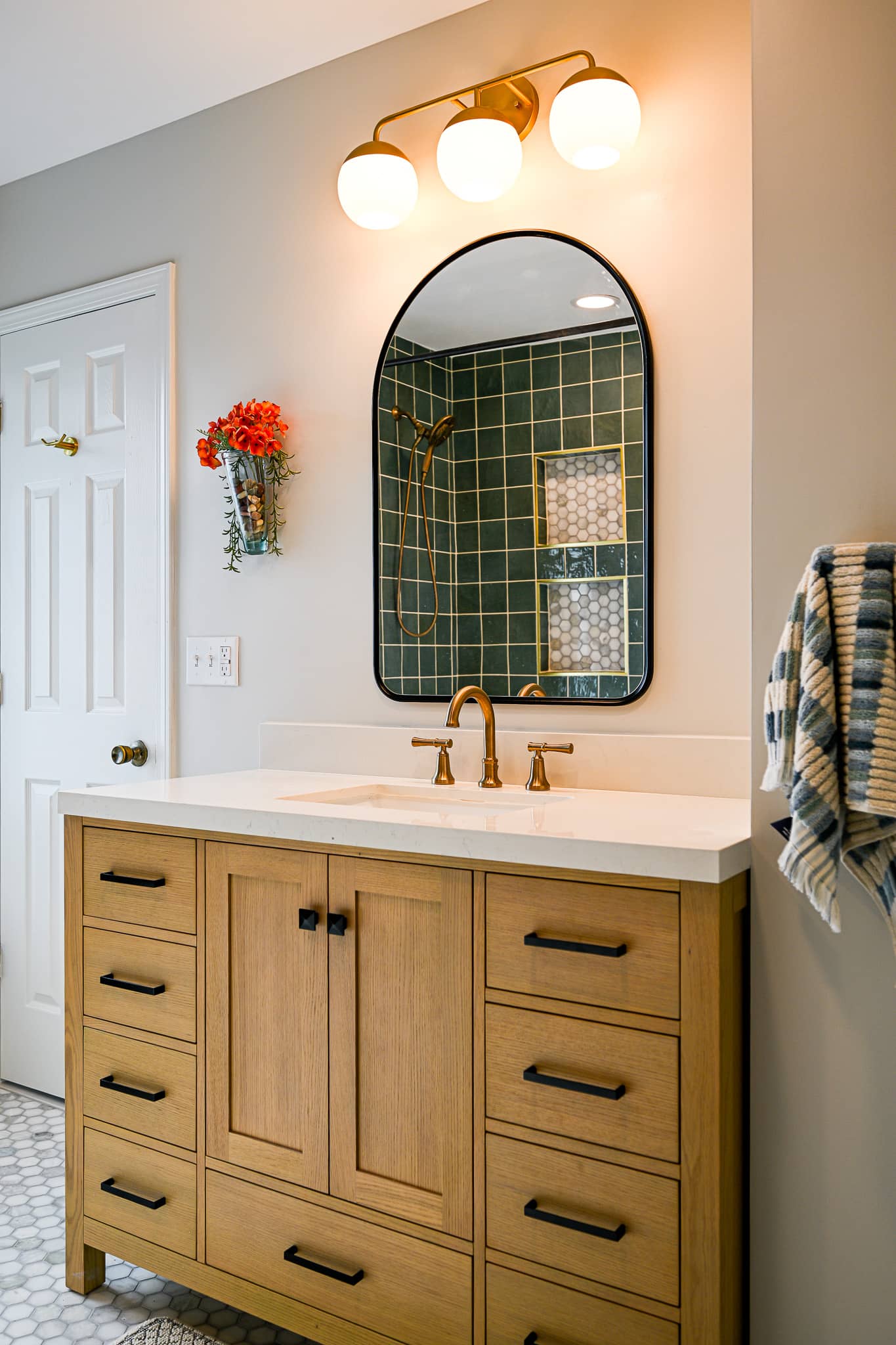 White oak vanity in the Dexter bathroom with black hardware, a brass faucet and an arched black framed mirror