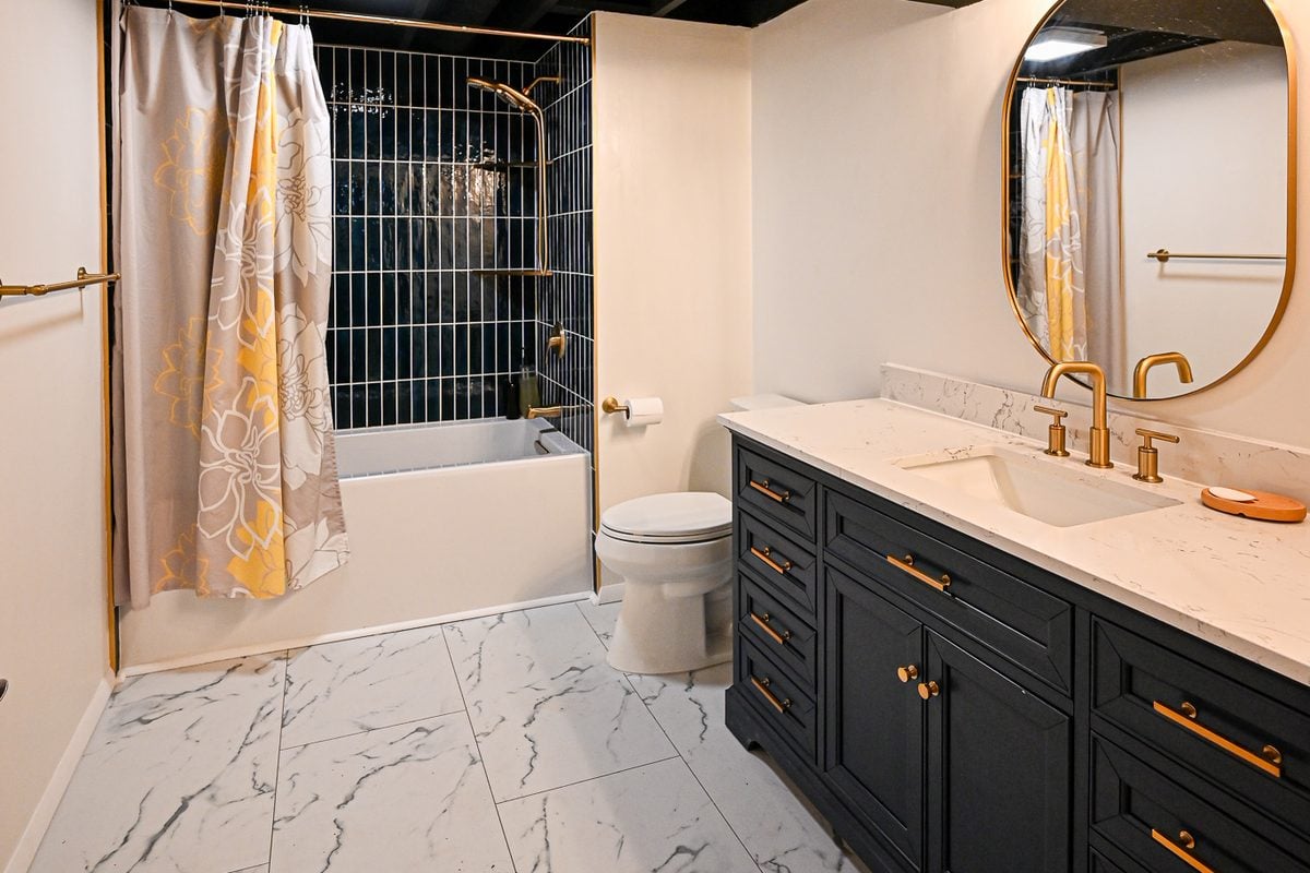 Finished basement bathroom in Ann Arbor with a navy tiled tub surround, a navy vanity with brass fittings and marble-look tile flooring