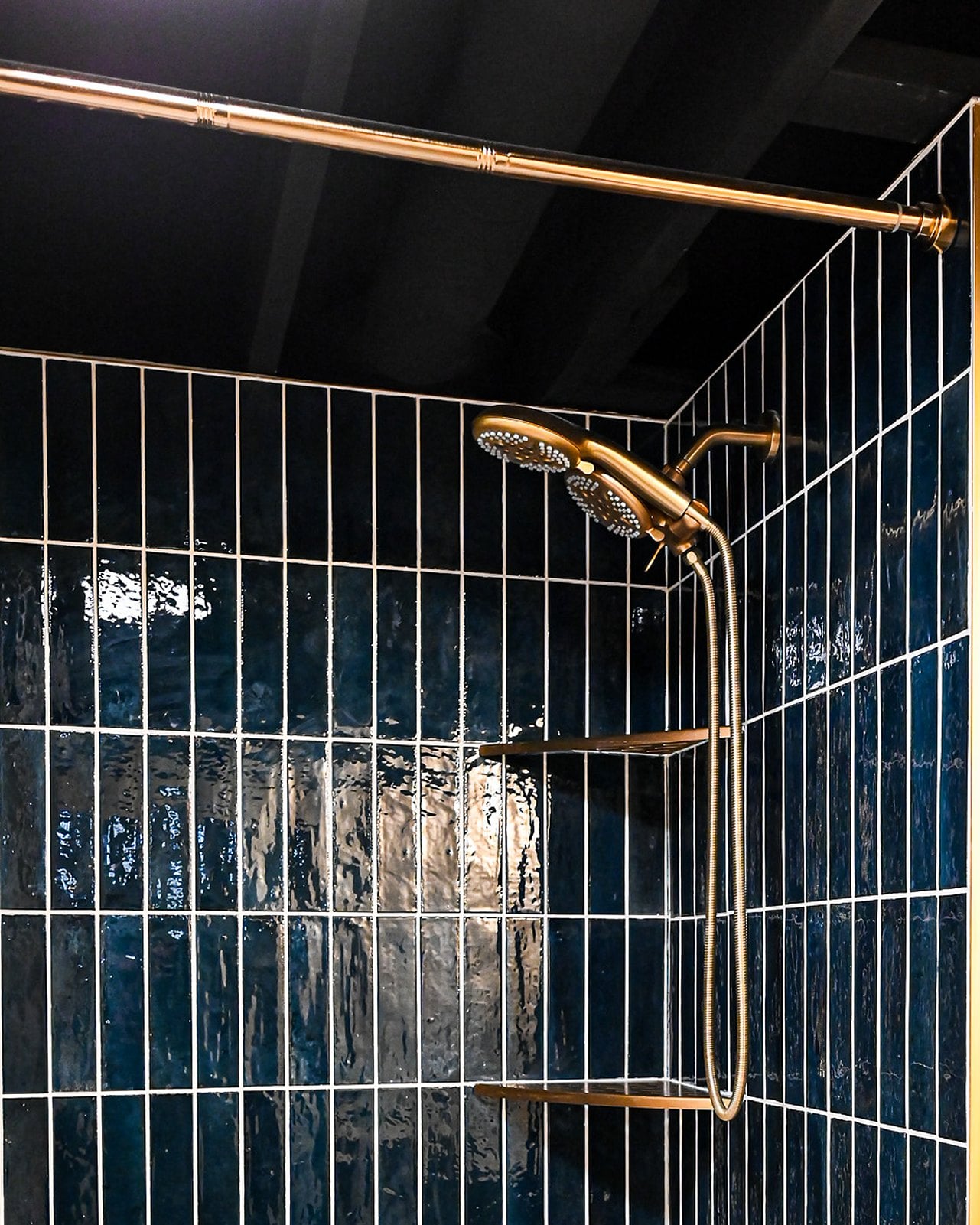 Navy stacked tile shower wall with two brass shower heads, a handheld on a slide bar and two brass corner shelves