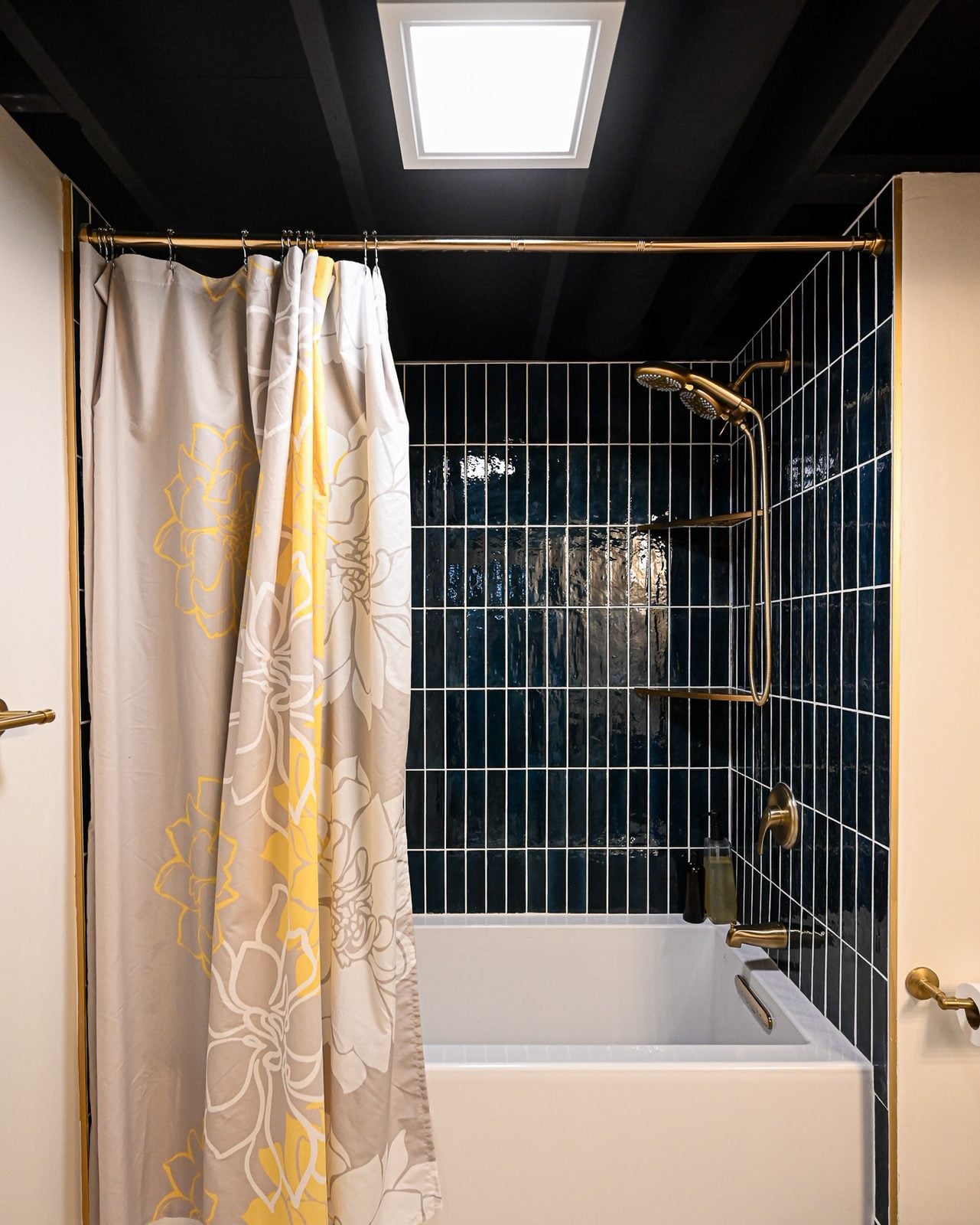 Alcove tub with a navy stacked tile surround, brass handheld shower and brass tub filler in a finished basement bathroom