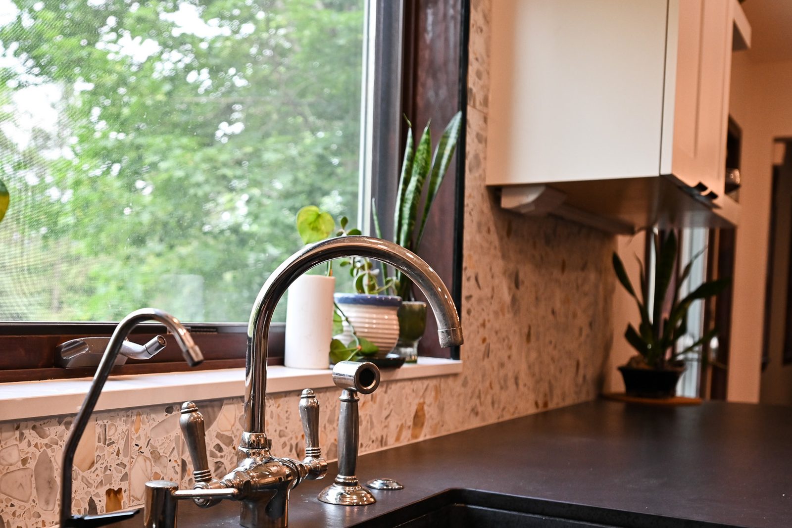 Polished nickel bridge faucet and black composite apron front sink below a window against a terrazzo backsplash