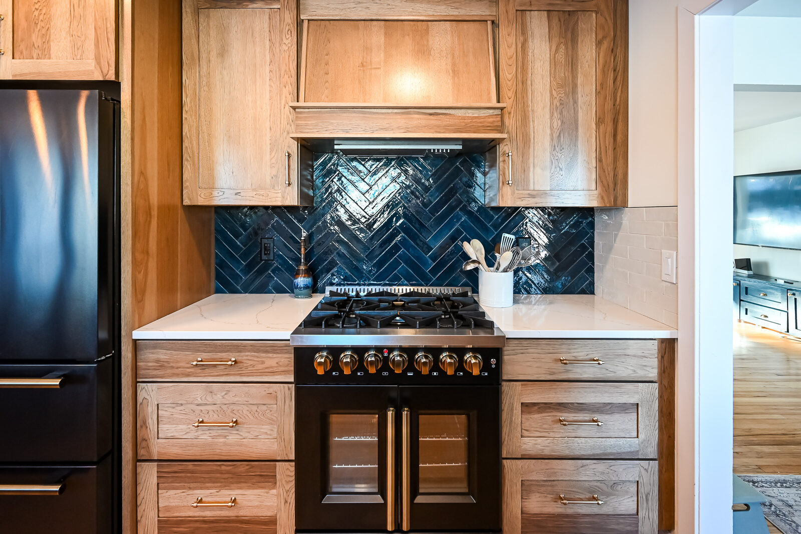 Glossy navy herringbone tile behind a black and brass range, framed by a wood hood surround and quartz counters