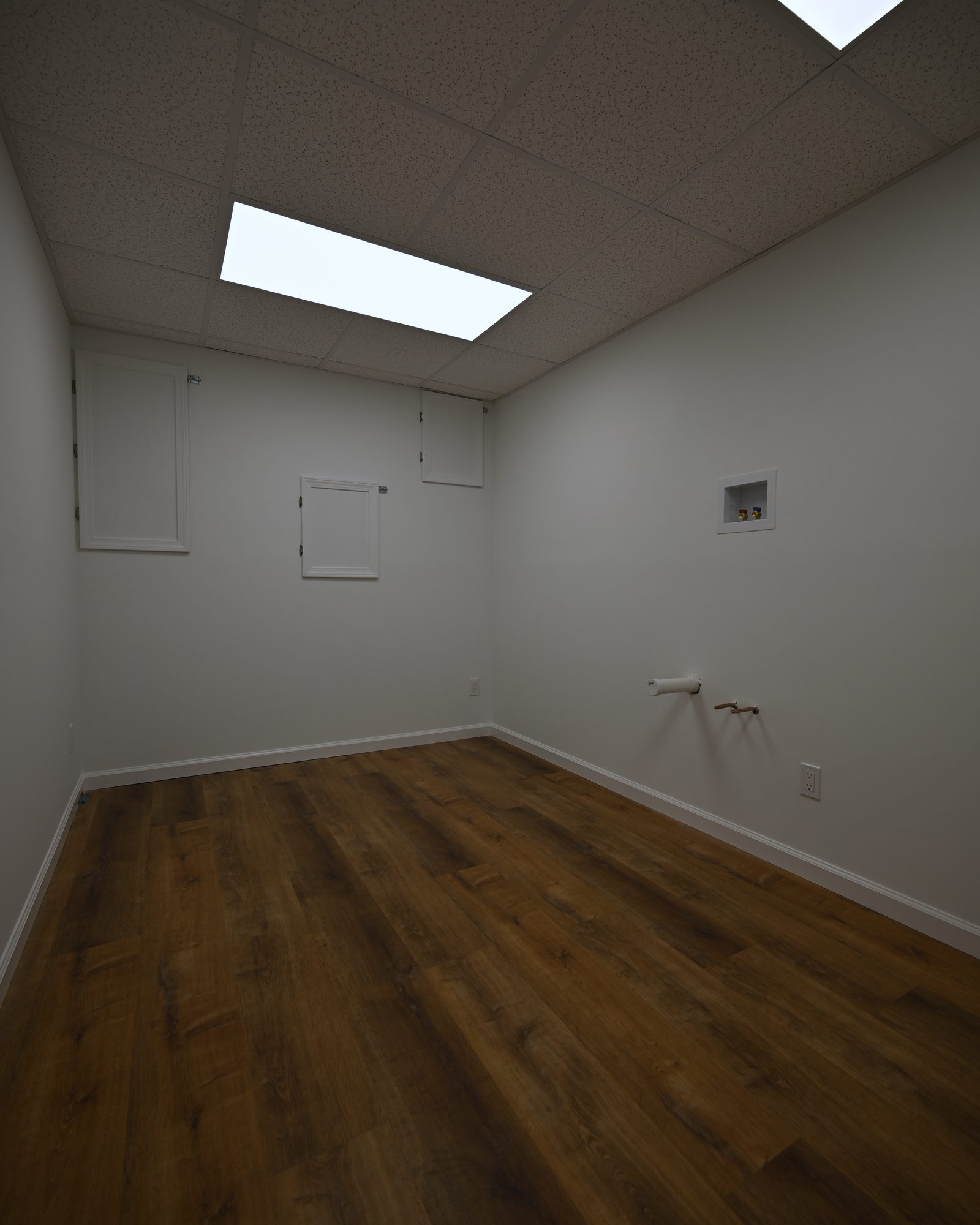 Darkroom wall with three flush access panels, a recessed supply box and a capped plumbing stub below a drop ceiling with LED flat panels