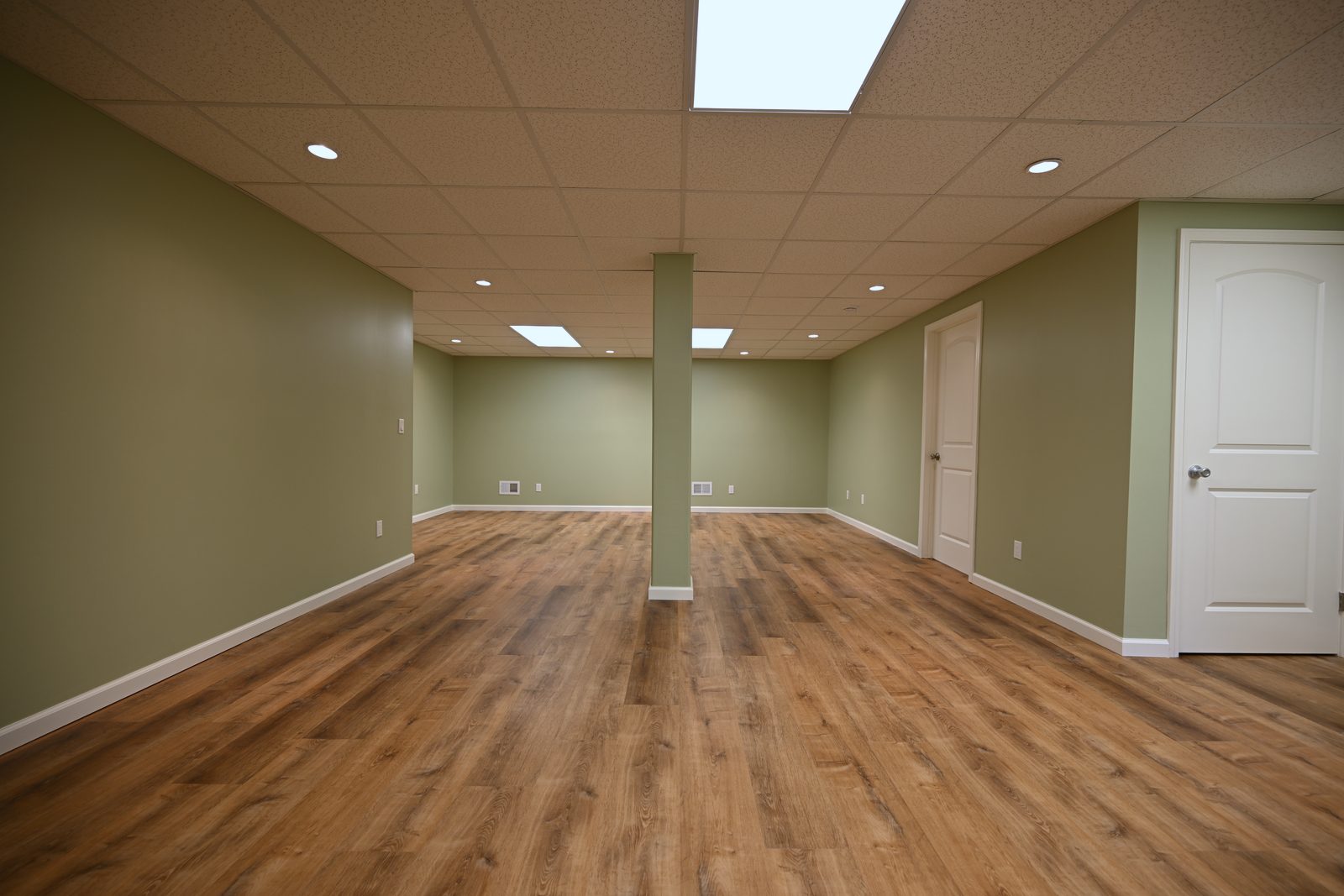 Long view of a finished basement rec room in Ann Arbor with a wrapped structural post, sage green walls and warm oak luxury vinyl plank floor