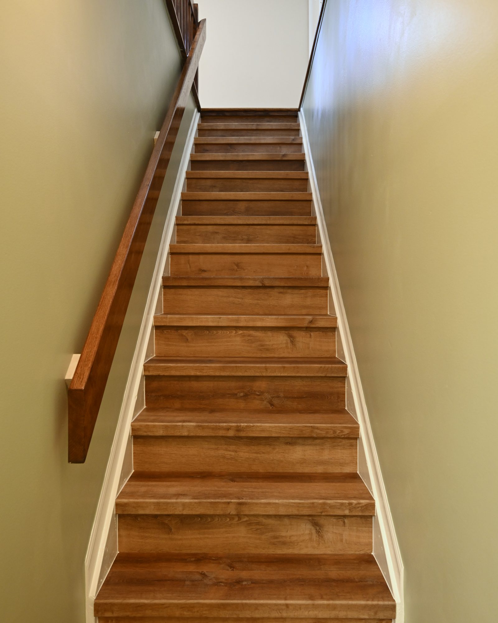 Basement stairway with luxury vinyl plank run over every tread, painted white risers and skirt board and a wood handrail
