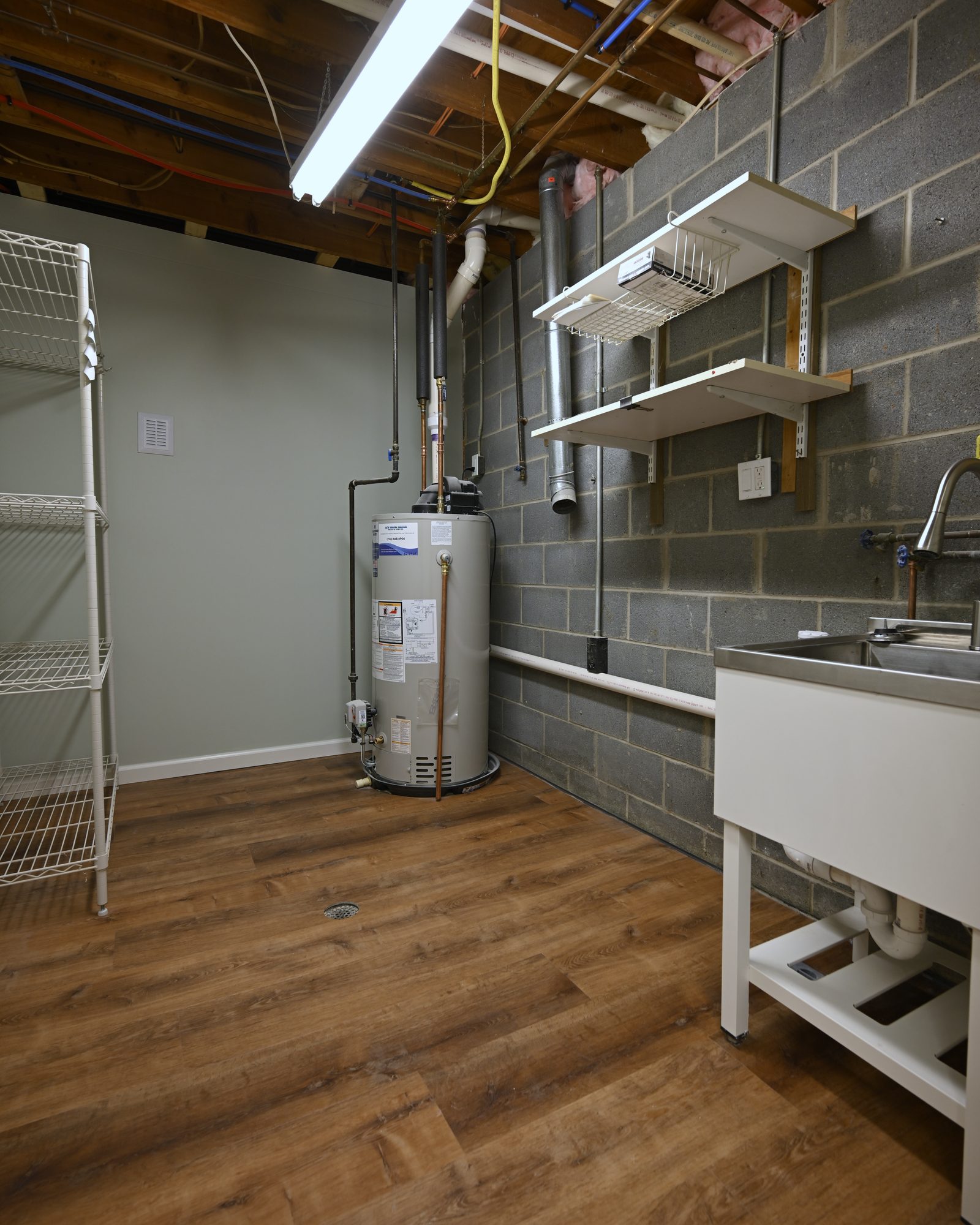 Basement utility room with exposed joists, a block wall, a gas water heater, wire shelving, a stainless utility sink and a floor drain set into new vinyl plank