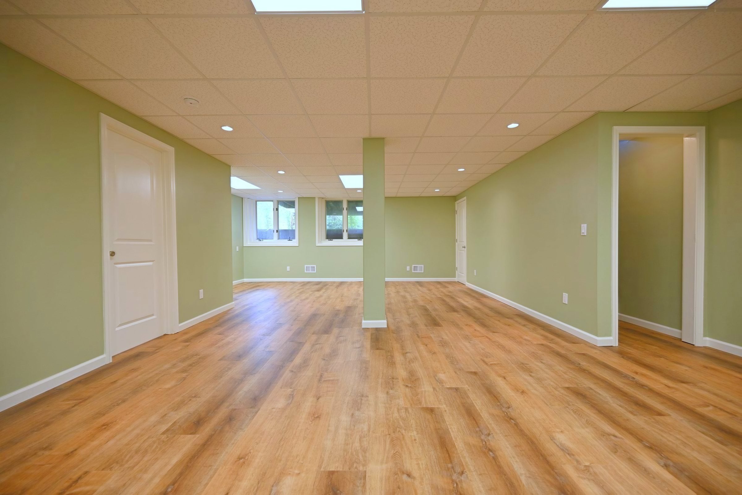 Finished basement room with two daylight windows, drywall returns at the openings, sage walls and a doorway through to a hallway