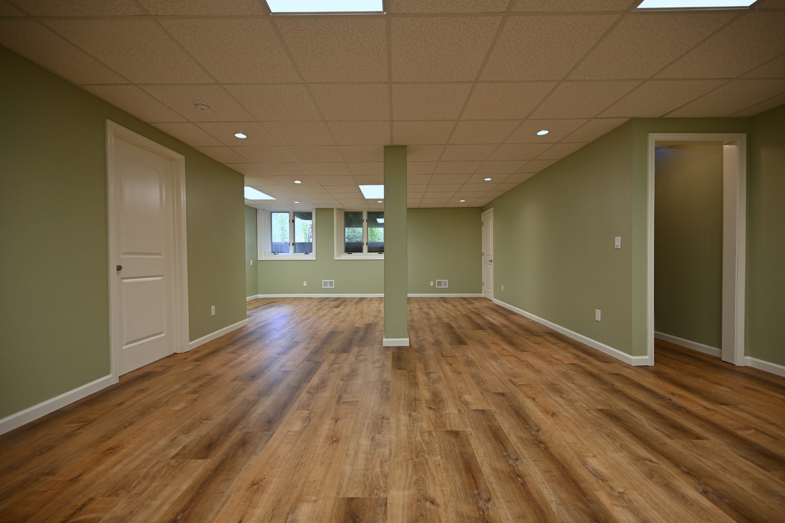Finished basement room with two daylight windows, drywall returns at the openings, sage walls and a doorway through to a hallway