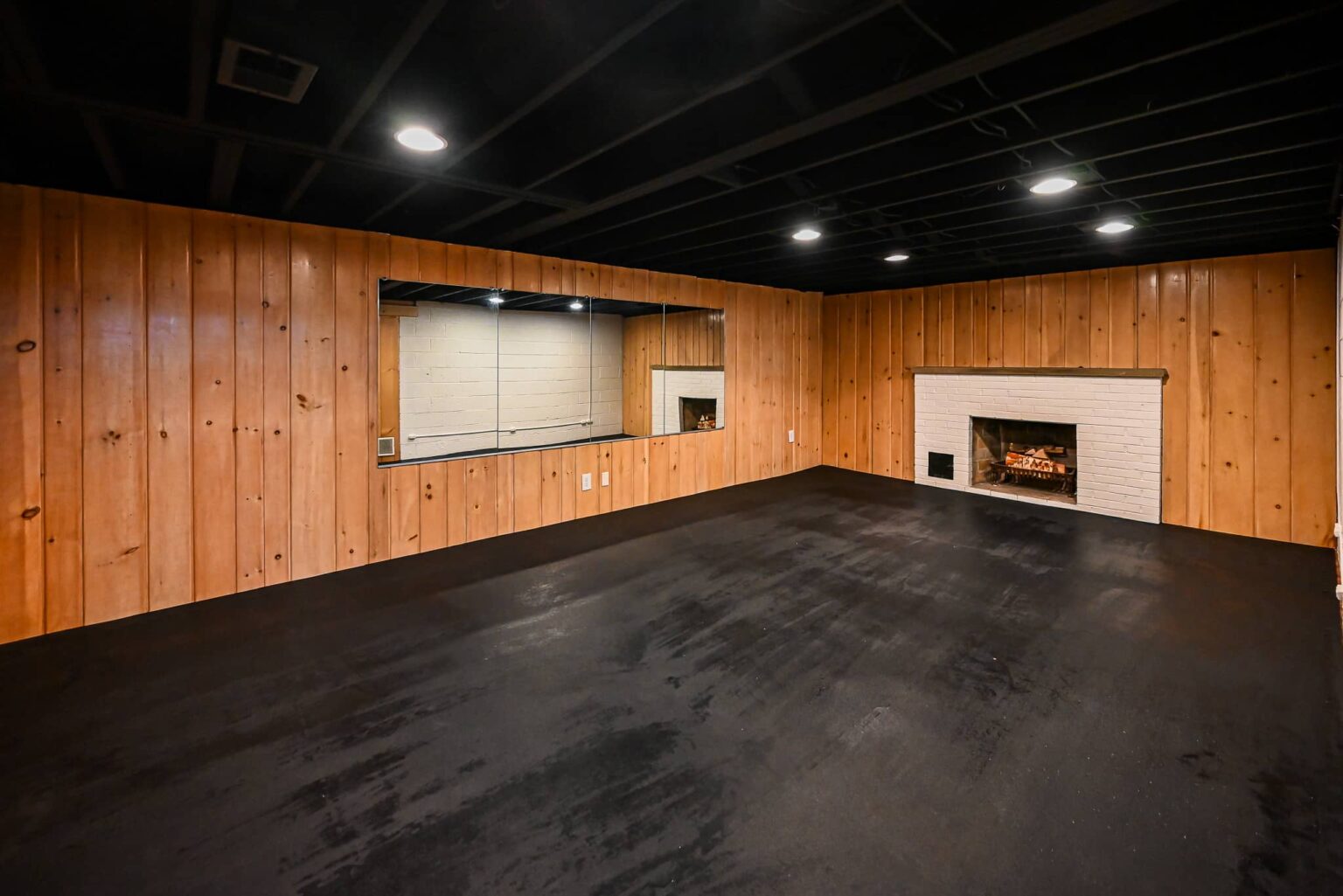 Basement gym wall with knotty pine paneling, full-length mirrors and a painted white brick fireplace