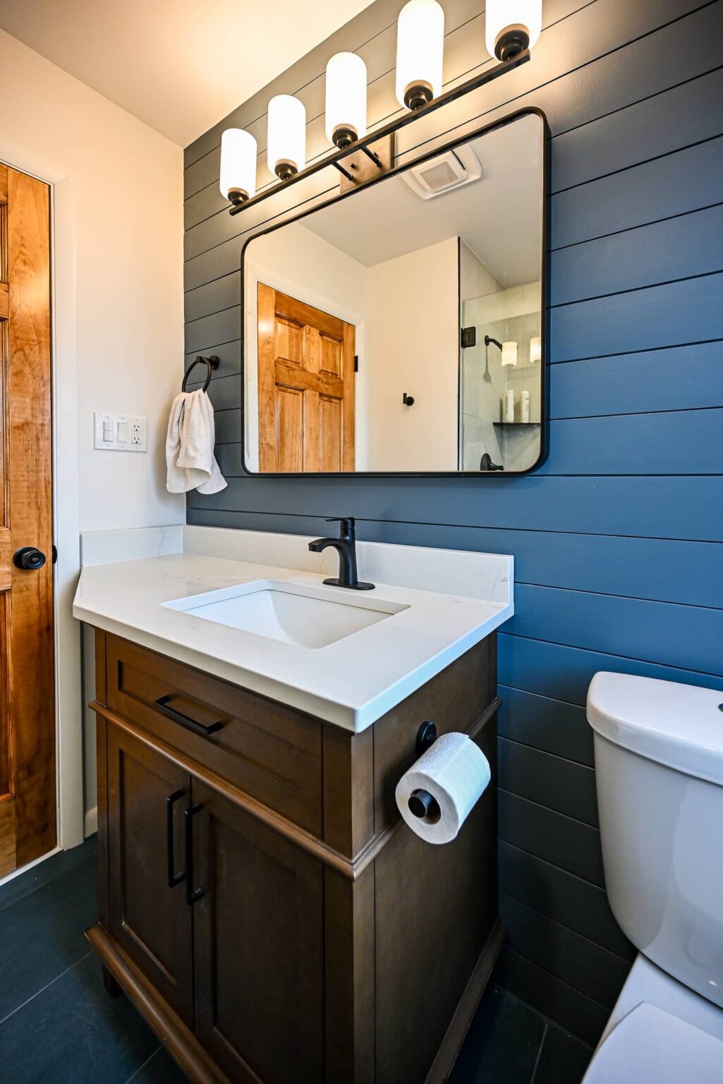 Dark wood bathroom vanity with a white quartz top, matte black faucet and black-framed mirror against navy shiplap