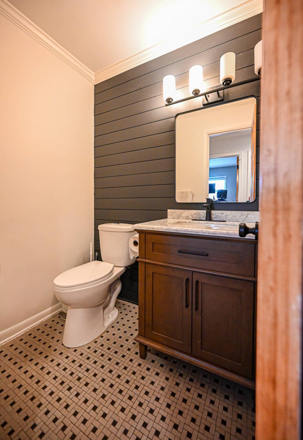 Half bathroom with a charcoal shiplap wall, chair rail, dark wood vanity with marble top and a black and white patterned tile floor