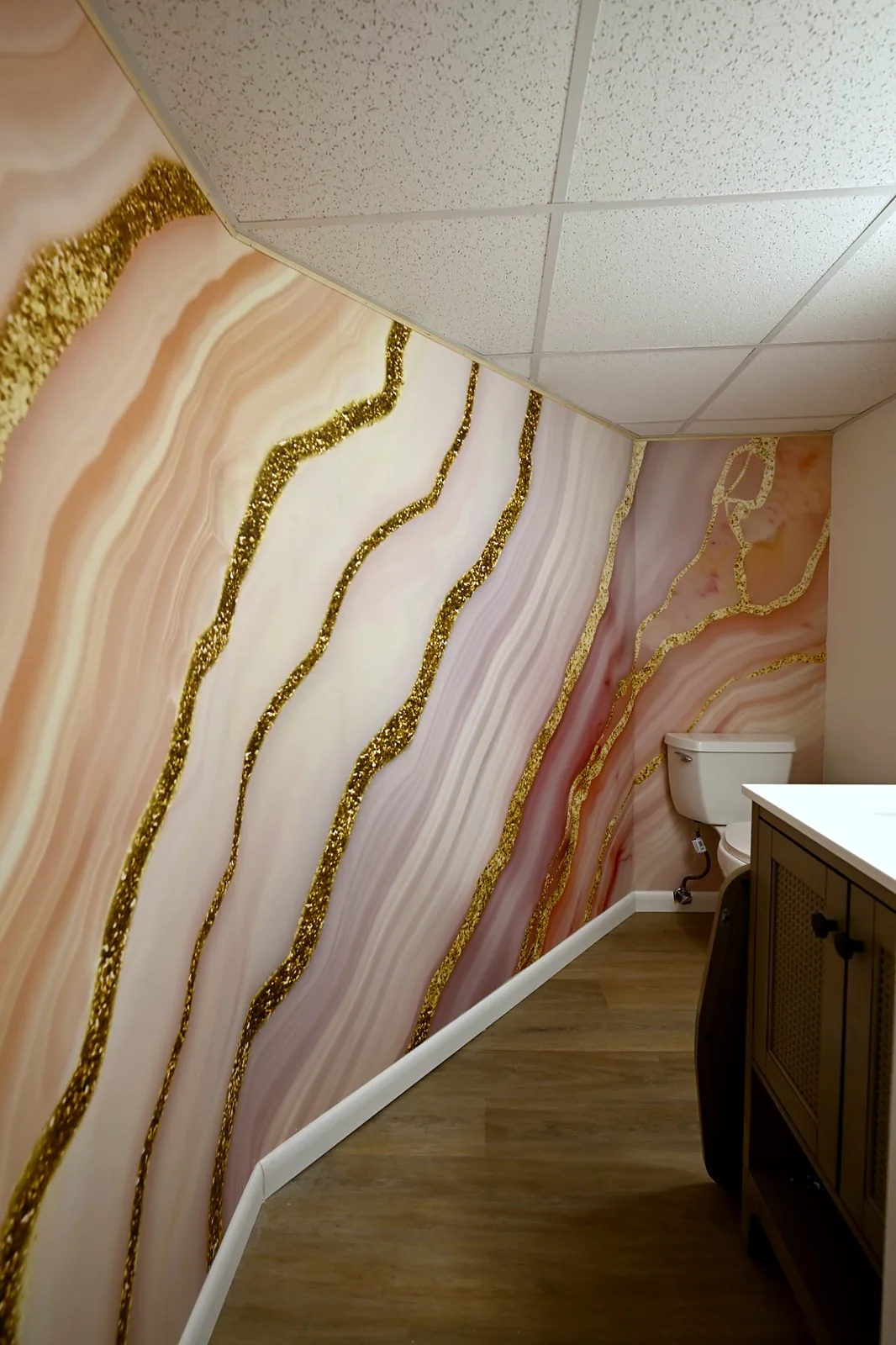 Basement powder room with a pink and gold agate wall mural running under a sloped drop ceiling, wood-look plank floor and a dark vanity