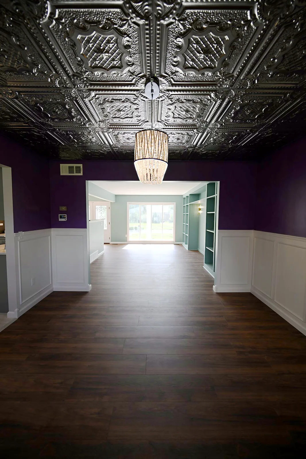 View from a purple dining room under a pressed metal ceiling through a wide opening to a teal great room with built-in shelving