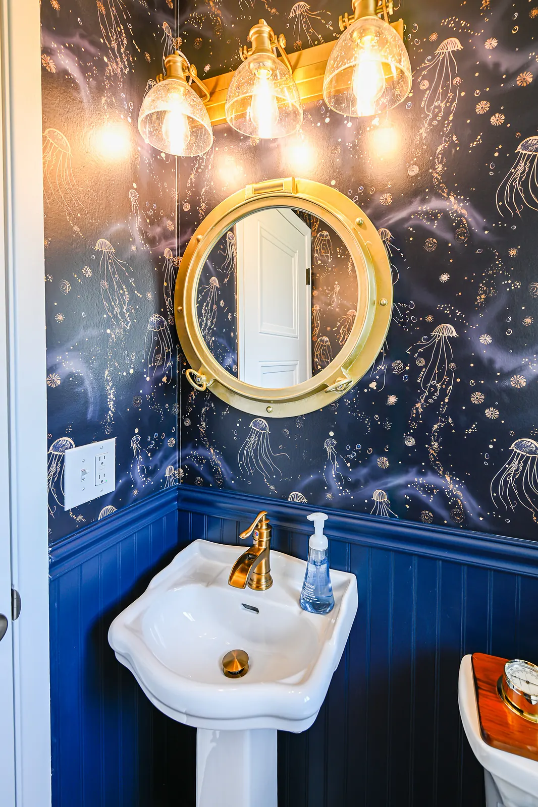 Pedestal sink with a brushed brass faucet under a brass porthole mirror and three-light seeded glass vanity fixture