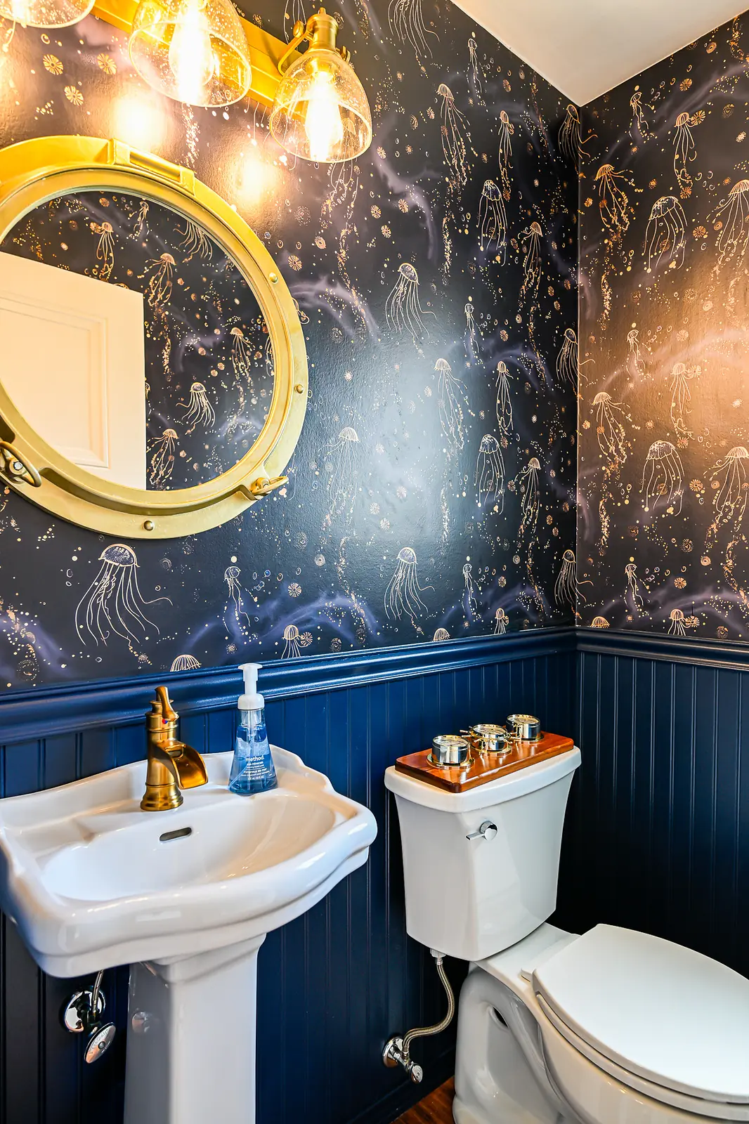 Powder room with navy jellyfish wallpaper above navy beadboard, a brass porthole mirror, brass vanity light and a white pedestal sink