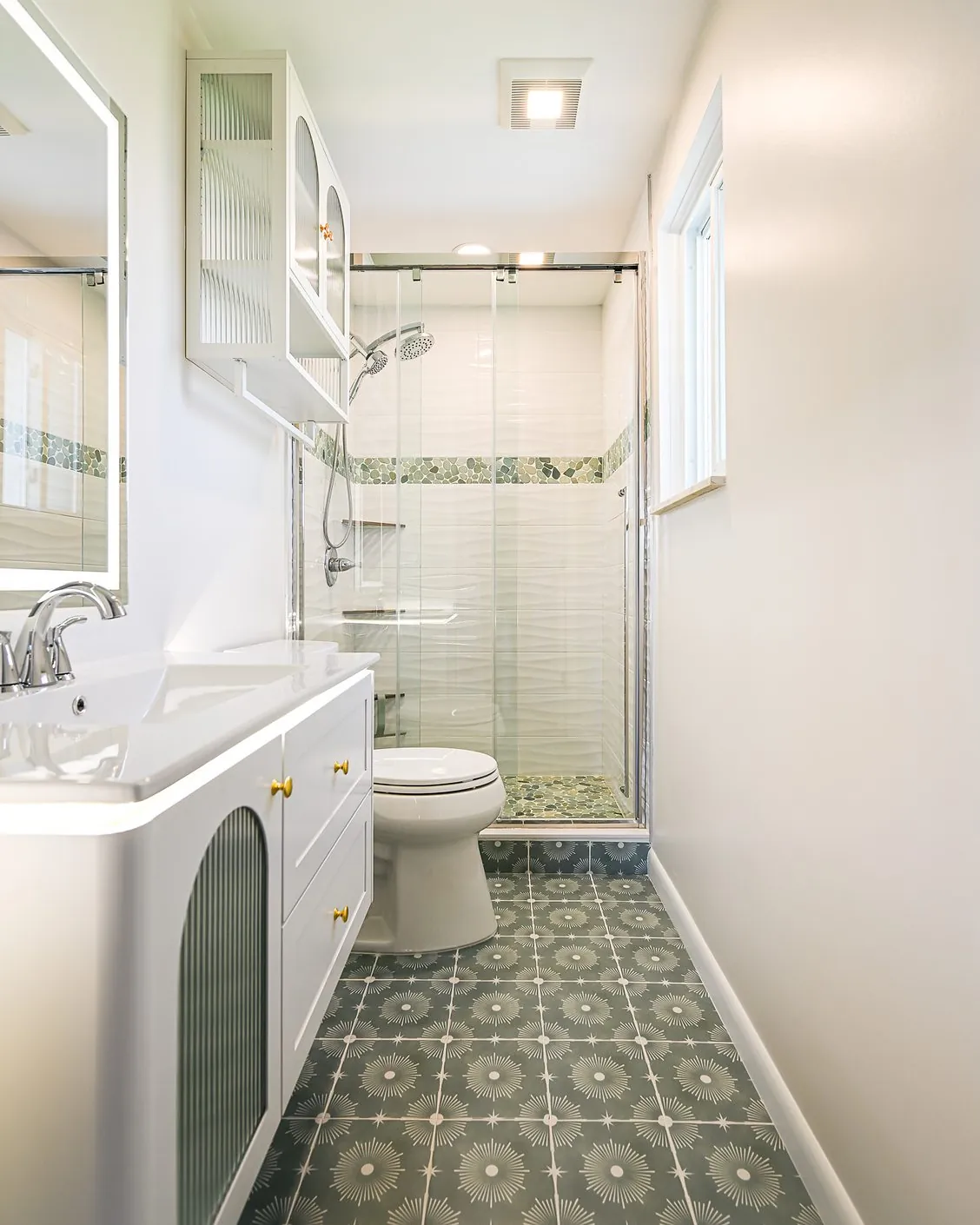 Narrow bathroom with a white floating vanity, green starburst cement tile floor and a glass-enclosed shower with a river pebble accent band