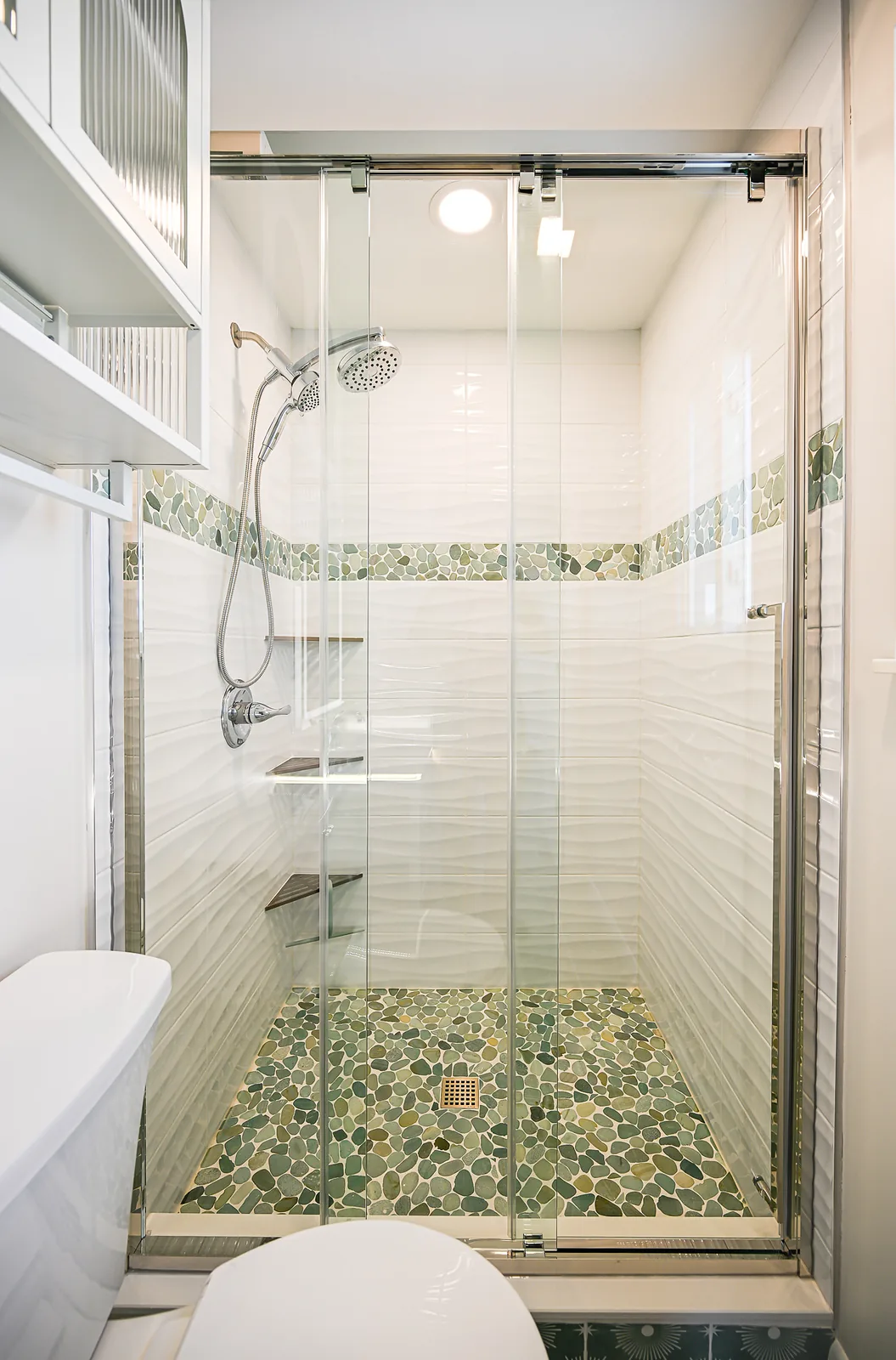 Walk-in shower behind frameless sliding glass doors, with white wave tile, a green river pebble band and a pebble floor