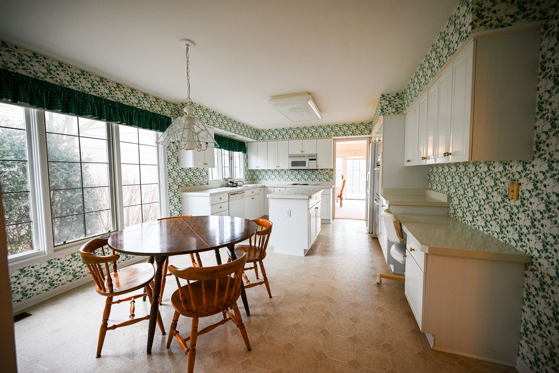 Kitchen before remodeling, with ivy print wallpaper, soffits above white cabinets and a vinyl floor