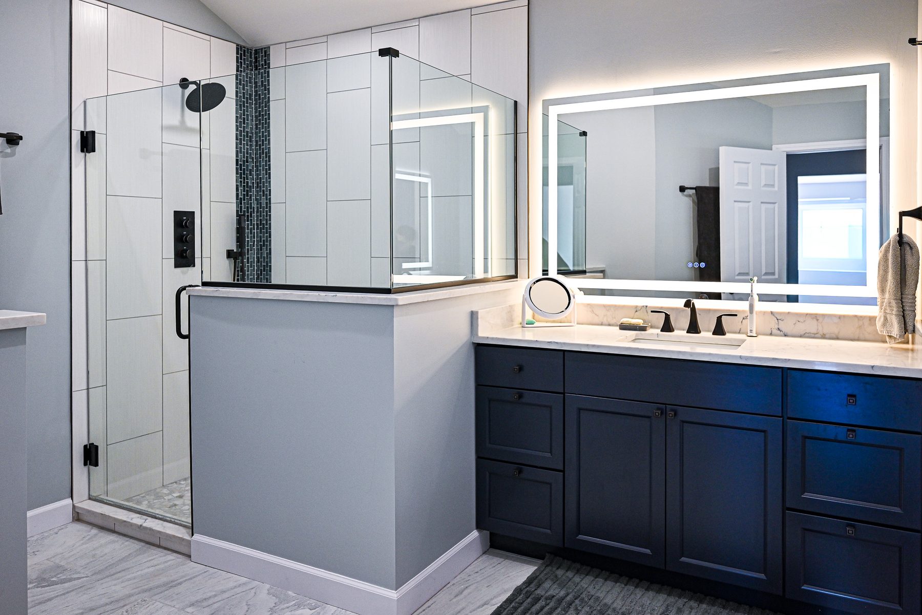 Renovated master bathroom with a frameless glass shower, backlit mirror, navy vanity and light tile floor