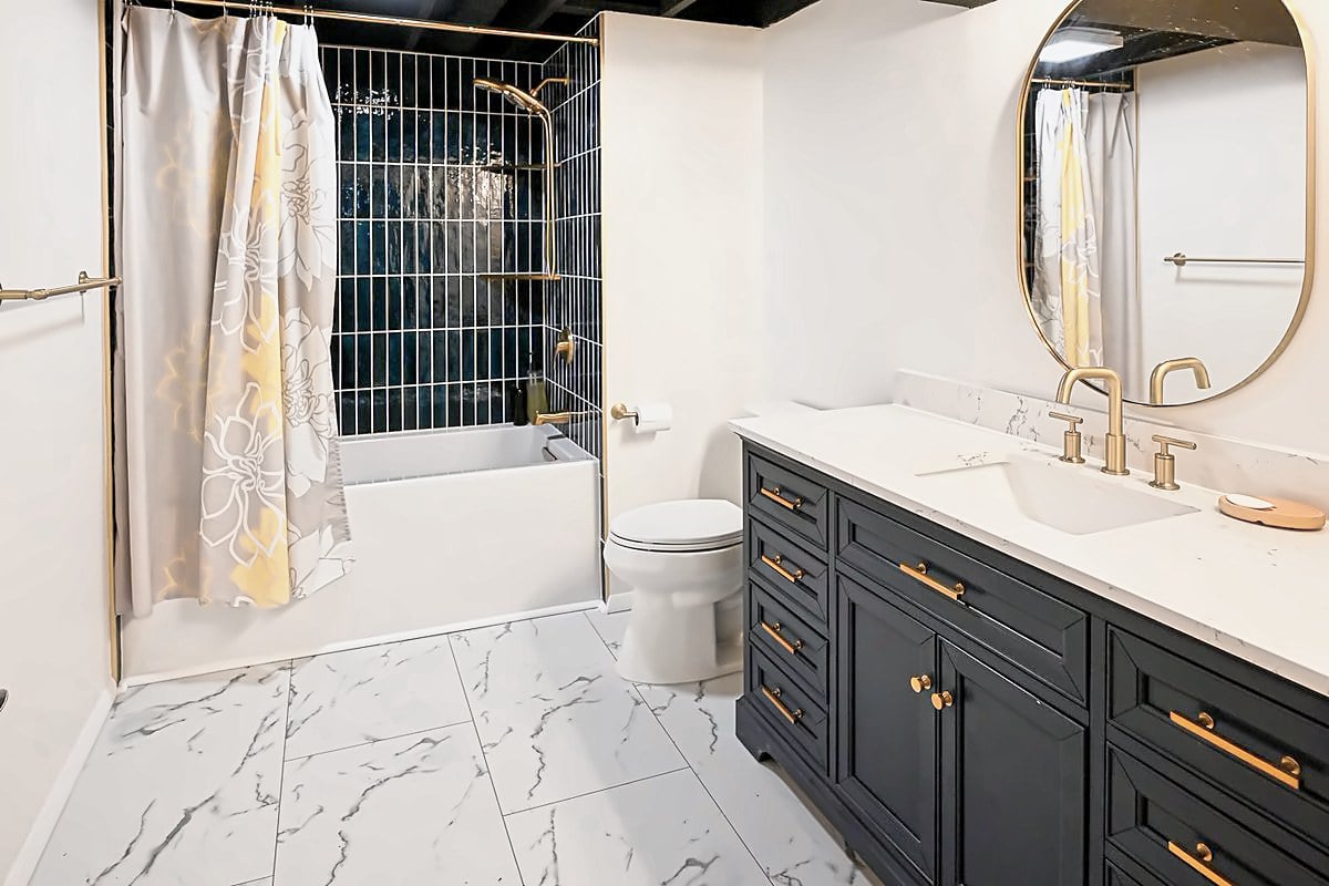 Finished basement bathroom in Ann Arbor with a navy tiled tub surround, a navy vanity with brass fittings and marble-look tile flooring
