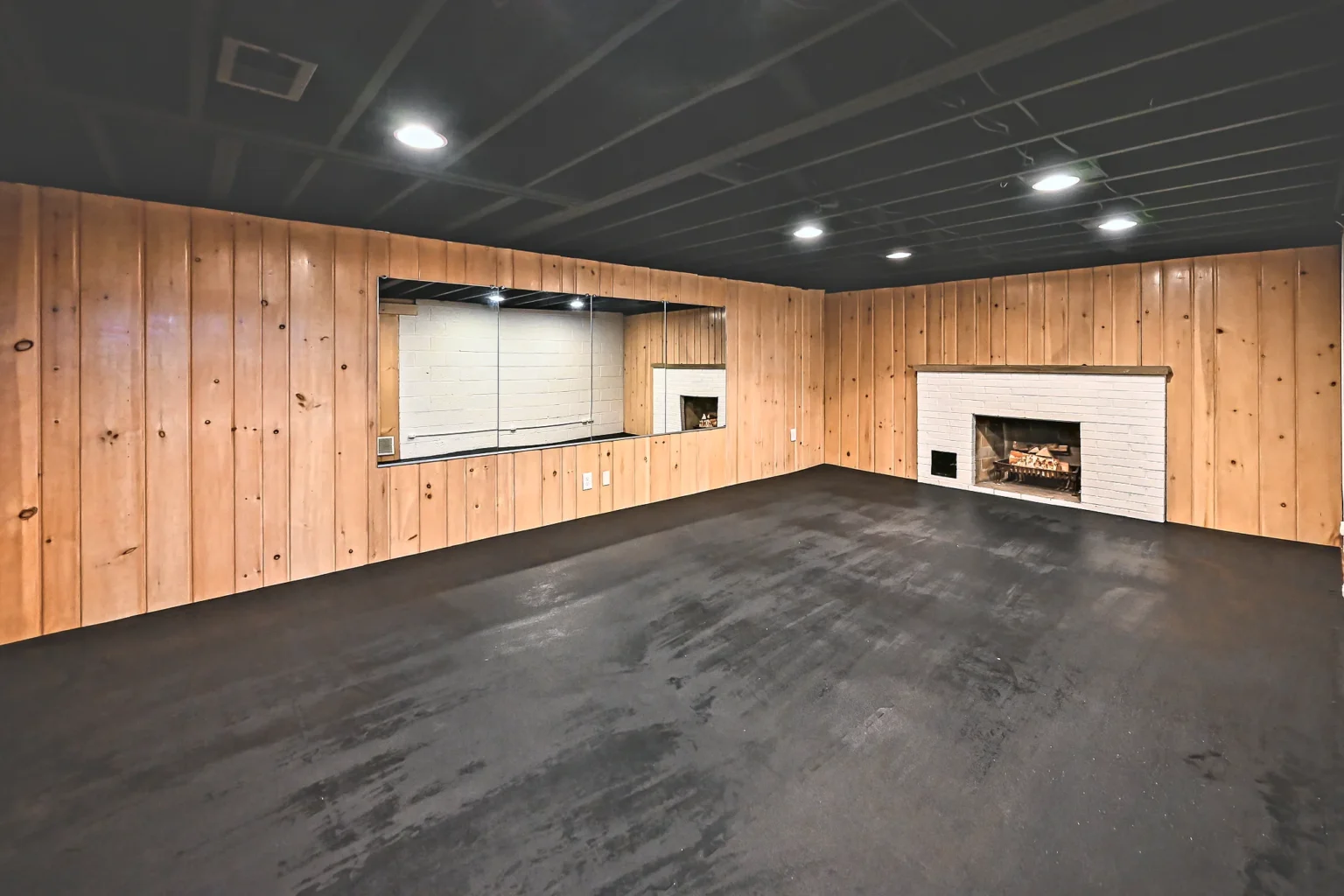 Basement gym wall with knotty pine paneling, full-length mirrors and a painted white brick fireplace