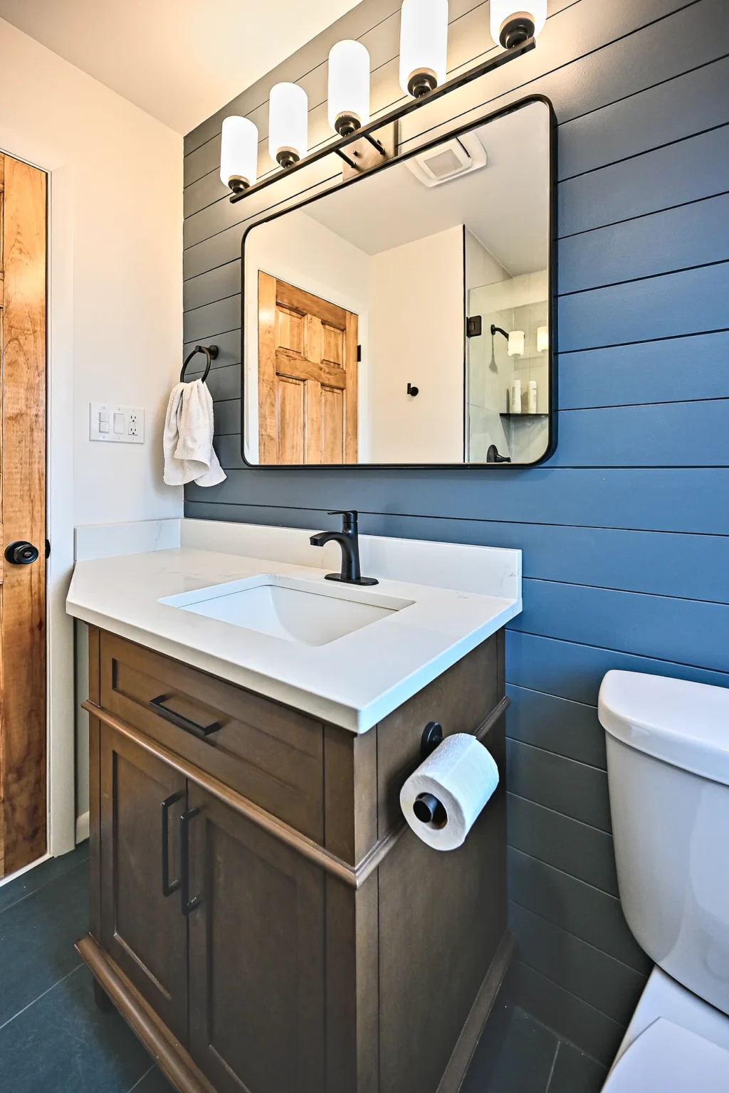Dark wood bathroom vanity with a white quartz top, matte black faucet and black-framed mirror against navy shiplap