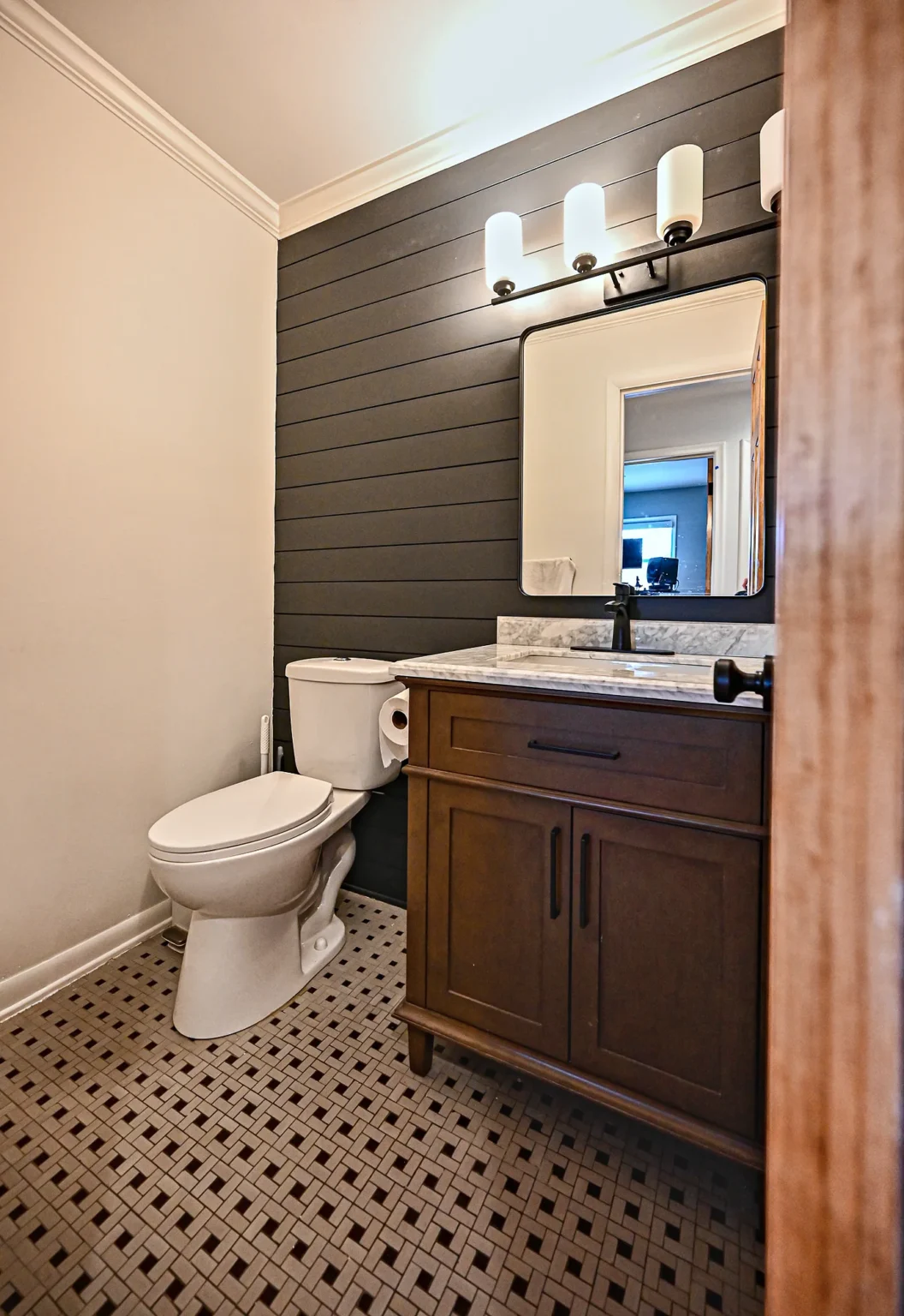Half bathroom with a charcoal shiplap wall, chair rail, dark wood vanity with marble top and a black and white patterned tile floor
