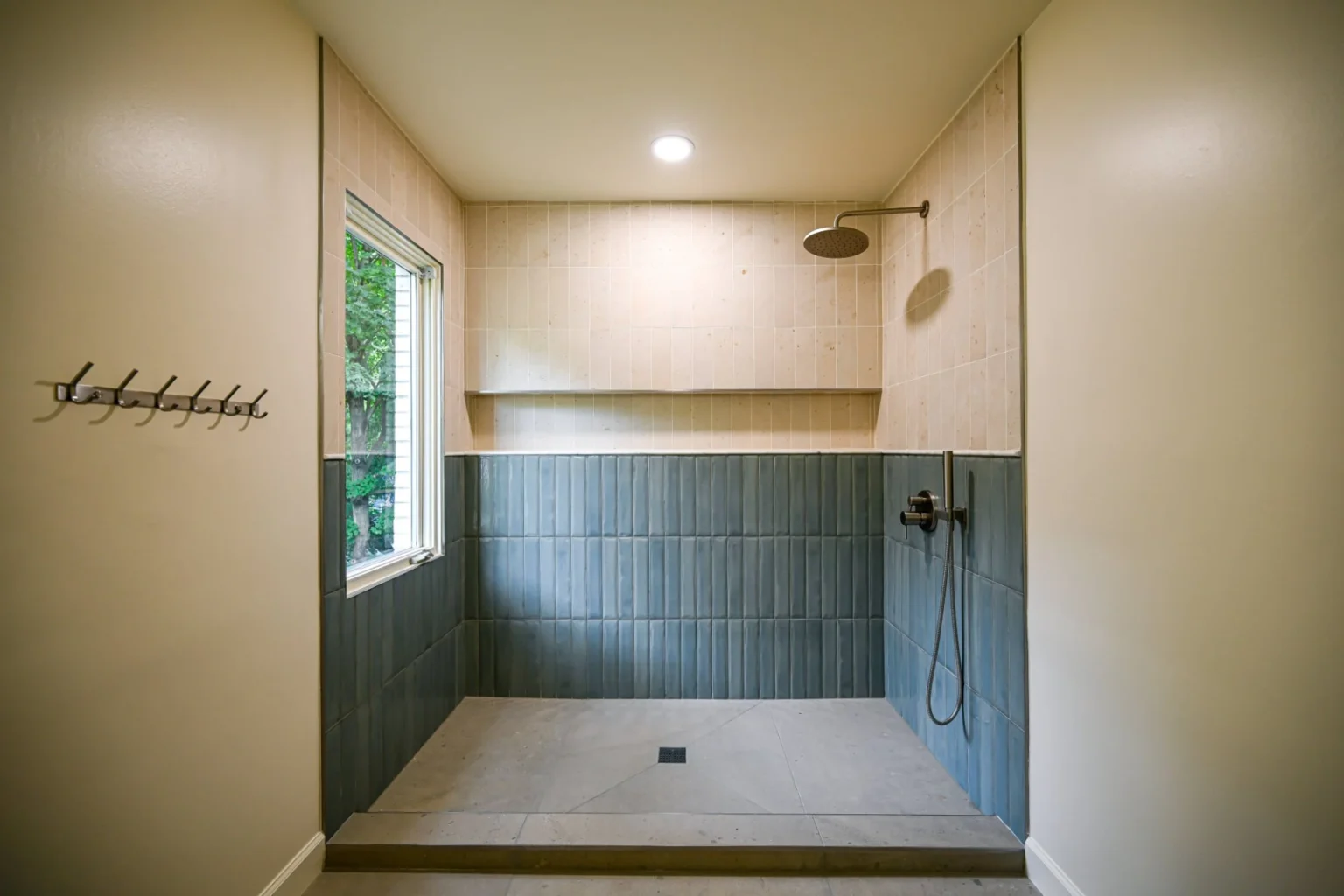 Walk-in shower in Ann Arbor with blue glazed tile below a limestone-look upper wall, a full-width ledge, rain head and a window to the trees