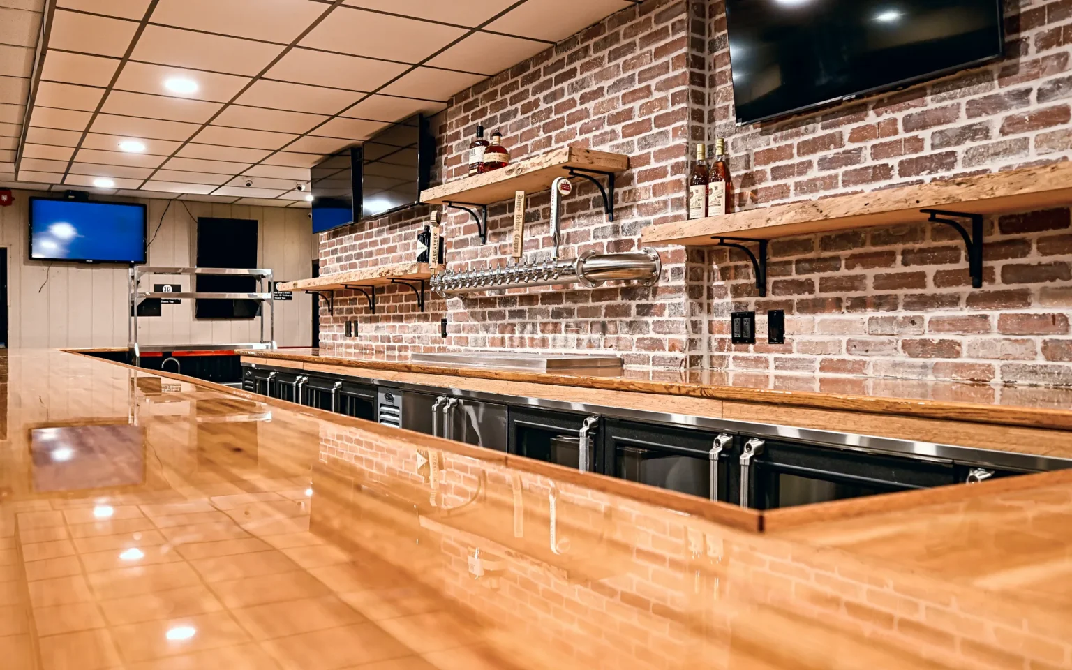 View along the butcher block bar top from the working side, showing the draft tower, back-bar coolers and live-edge shelves on brick