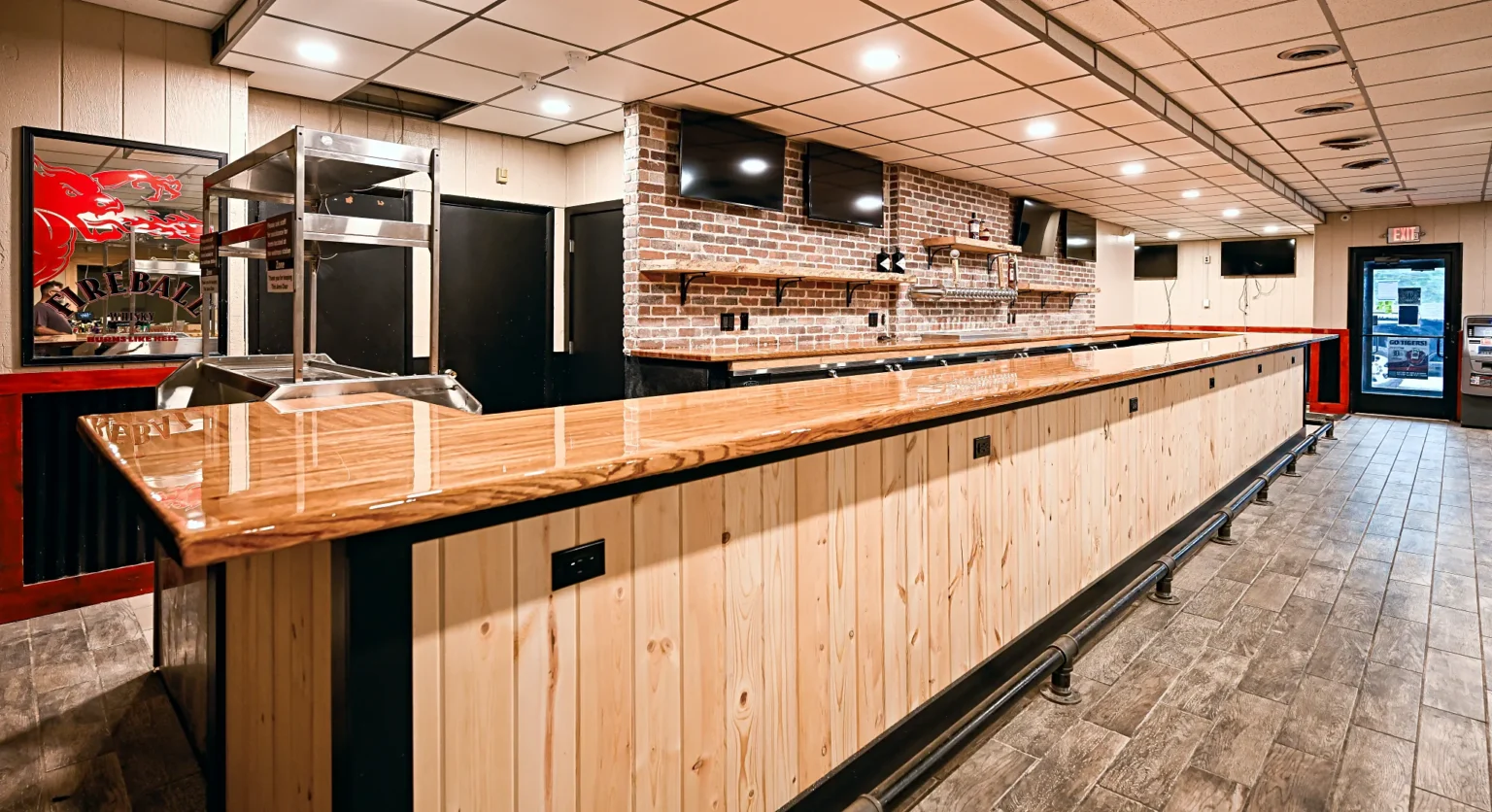 Corner of the new butcher block bar top turning toward the entrance, with the pine front, black steel band and the brick back bar beyond