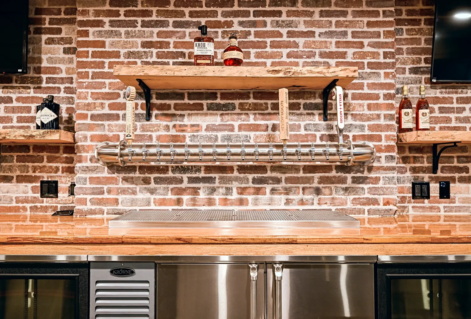 Stainless draft tower with a long row of taps mounted on a thin brick wall under a live-edge shelf, with a drip tray on the butcher block and back-bar coolers below