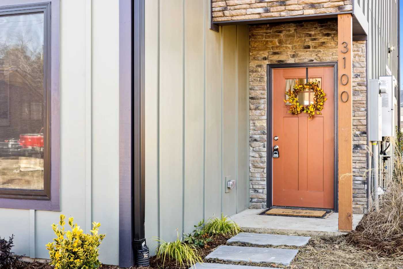 Orange front door with a fall wreath beside board and batten siding and a stone accent wall